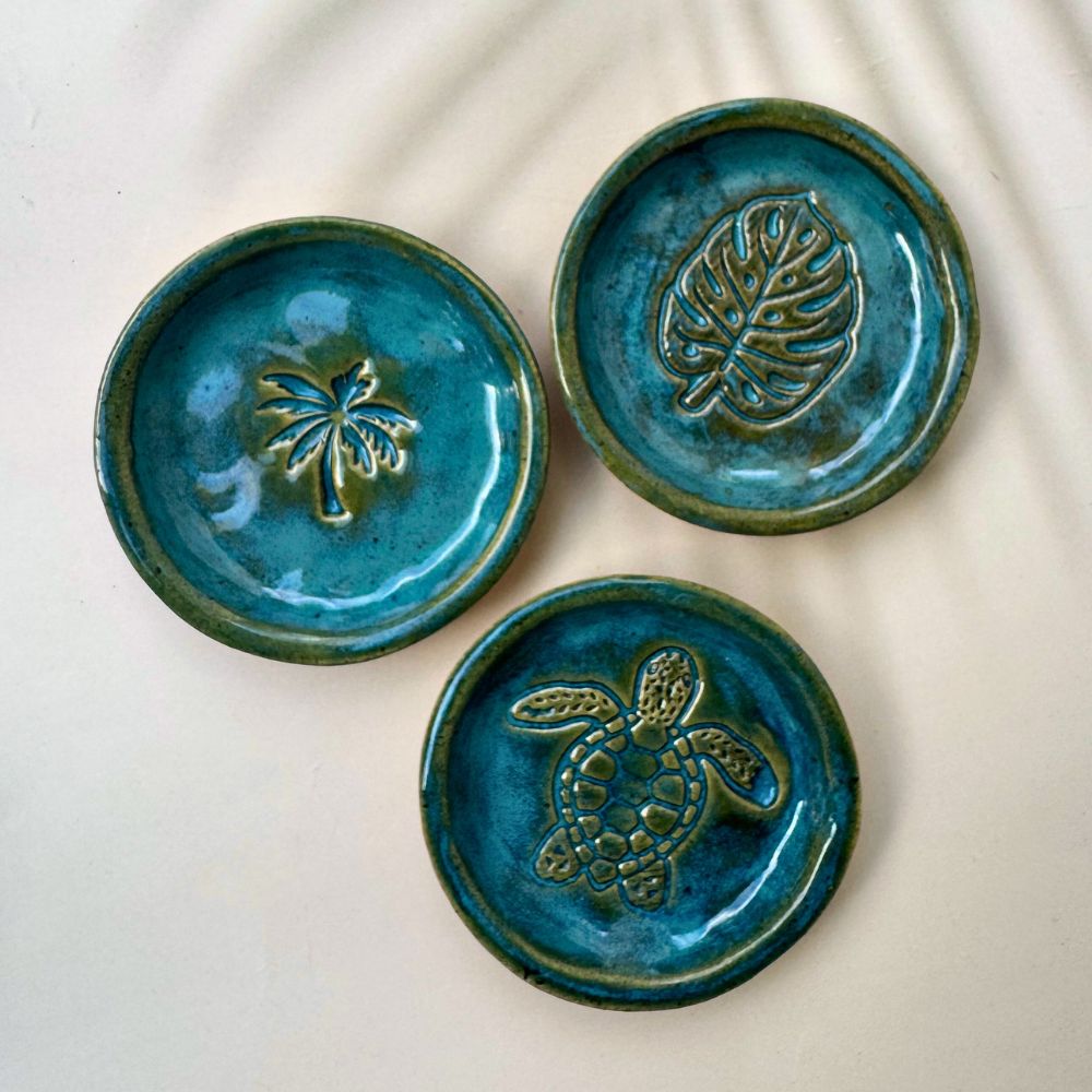Three blue ceramic coasters with embossed designs of a palm tree, leaf, and turtle on a white surface.