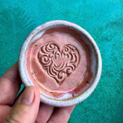 Hand holding a ceramic ring dish with a heart design on a teal background