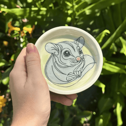 Hand holding a trinket dish with a sugar glider illustration against a green leafy background