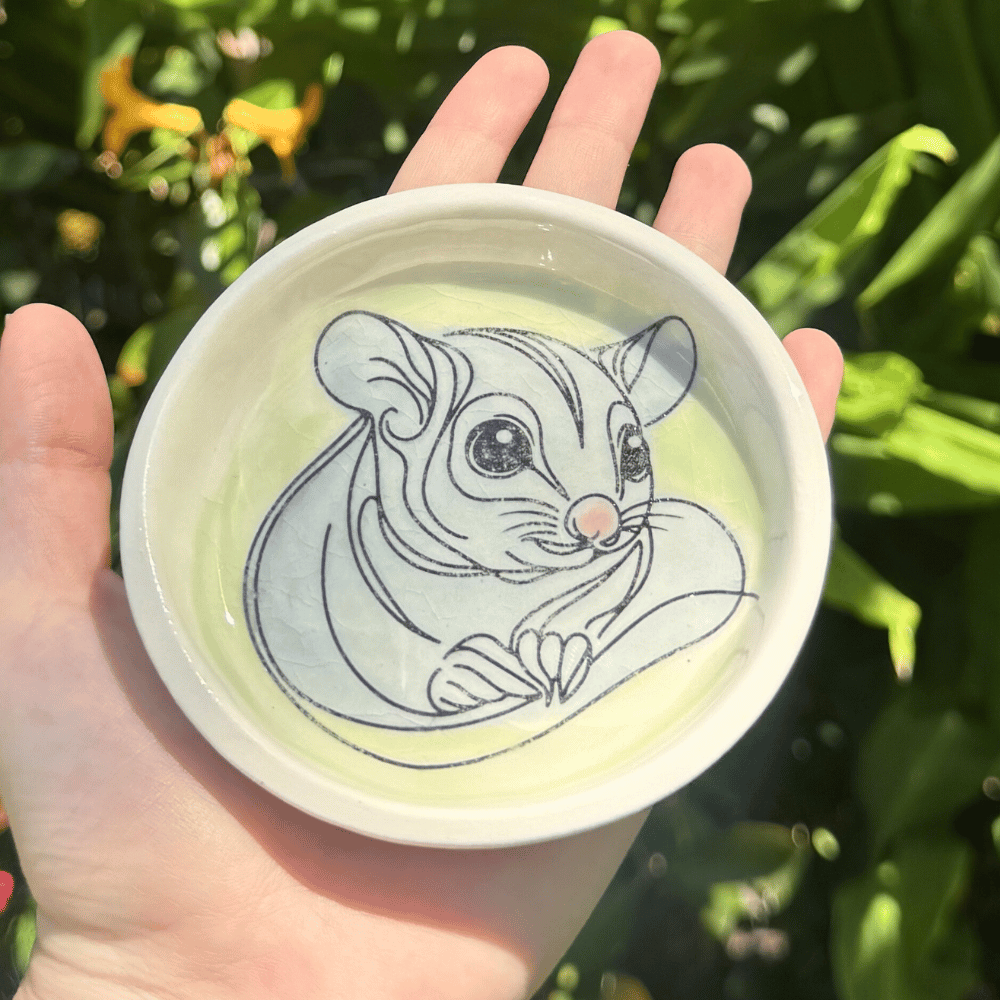 Hand holding a trinket dish with a sugar glider illustration against a green leafy background