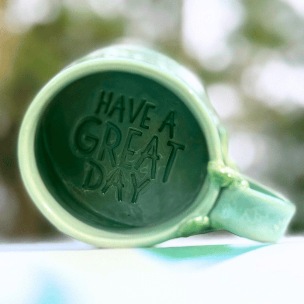 Green mug with 'Have a Great Day' text on a blurred natural background
