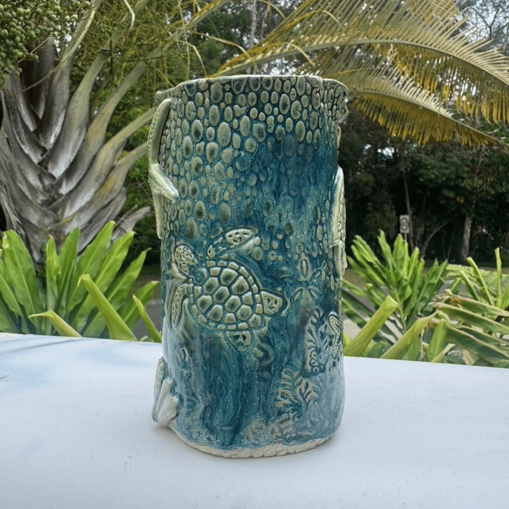 Blue ceramic vase with turtle design on a white surface with greenery in the background