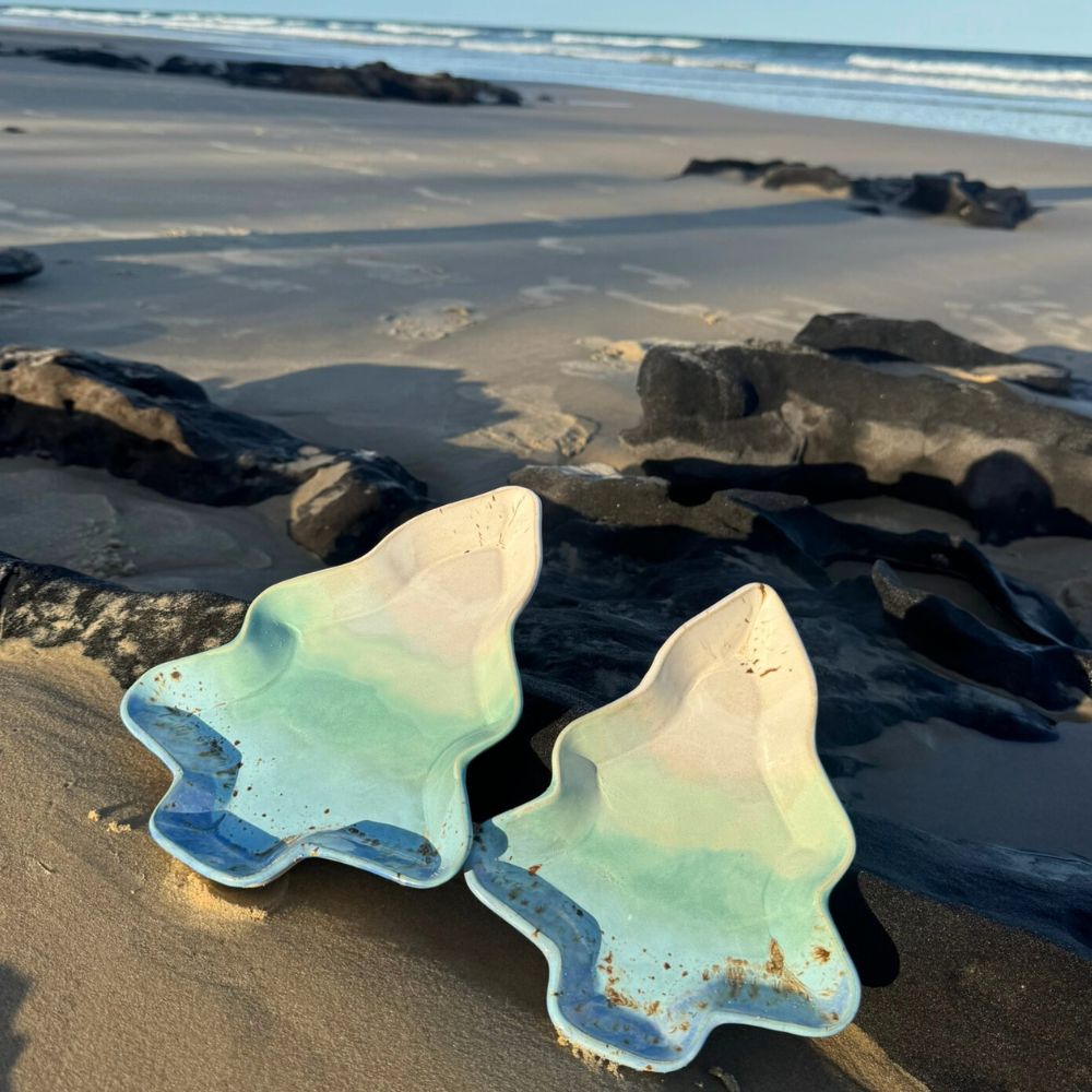 Two ceramic xmas tree plates on a sandy beach with rocks and ocean in the background.